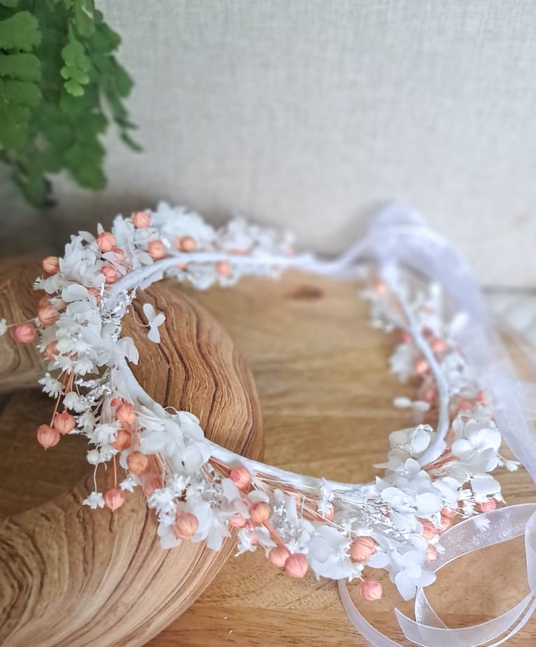 Delicate white and coral floral headpiece with dried flowers draped over a wooden head form on a wooden surface.