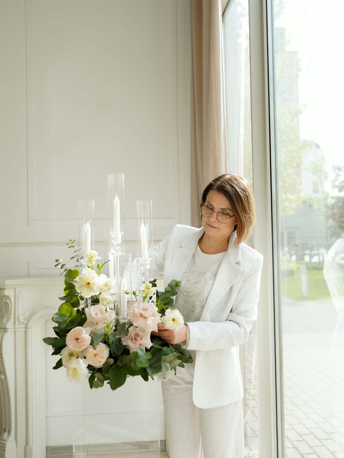 Woman in white blazer holding a vase with pink roses and white flowers with glass candles by a window