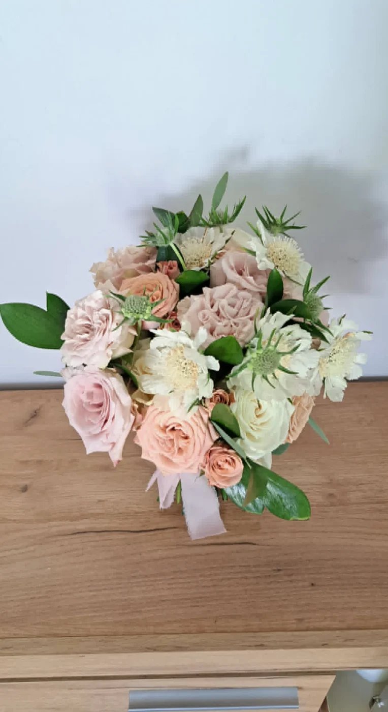 A pastel flower bouquet with blush pink roses, cream-colored flowers, and green foliage tied with a light pink ribbon, displayed on a wooden surface