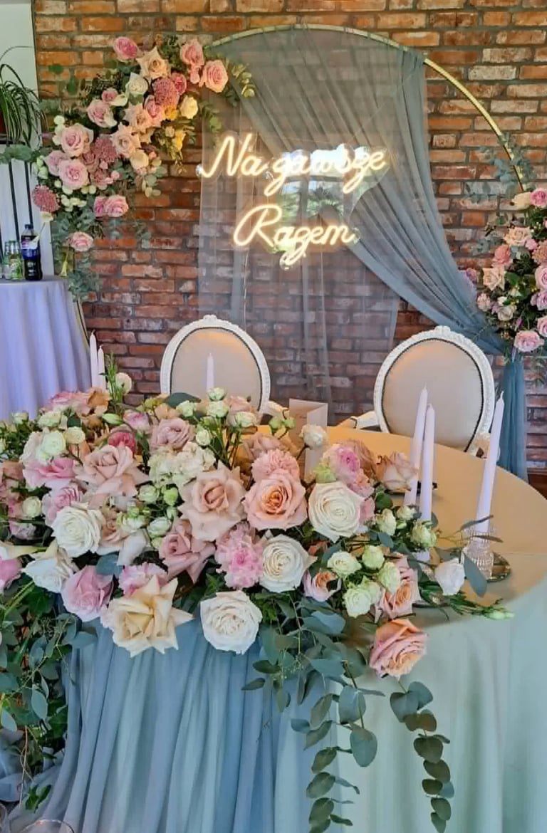 Elegant wedding reception sweetheart table with pink and cream floral arrangement, neon signage, and blue draping against brick wall