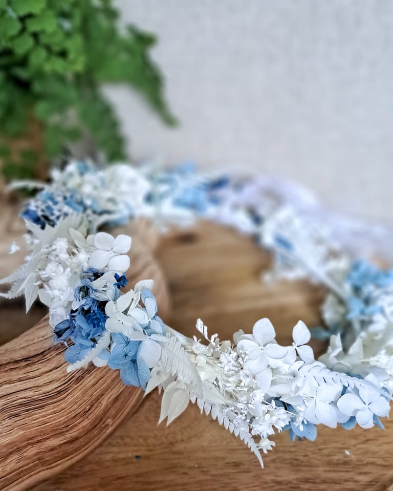 Floral wreath with white and blue dried flowers arranged on a wooden base with green foliage backdrop