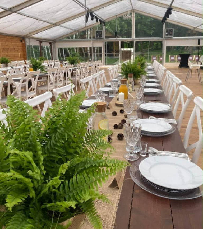 Elegant indoor dining setup in a glass pavilion with white chairs, long wooden tables set with place settings, green plants, and natural lighting from the arched ceiling