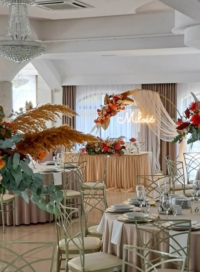 Elegant wedding reception venue with cream and gold decor, floral arrangements, neon sign reading More, and formally set dining tables