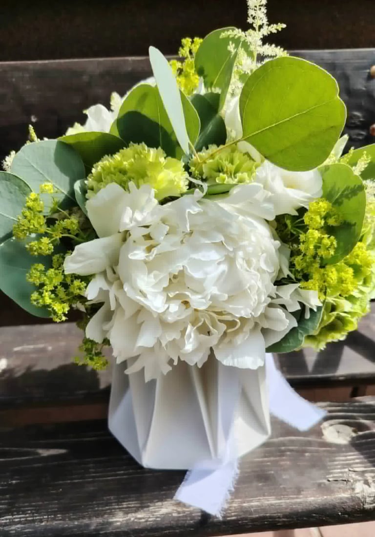 White floral arrangement with peonies, greenery, and yellow accents in a white vase with ribbon bow