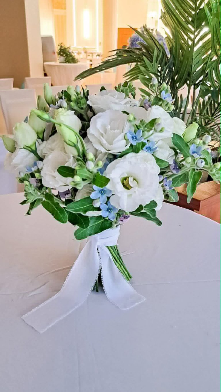 White bridal bouquet with roses, blue forget-me-nots, and green foliage tied with white ribbon