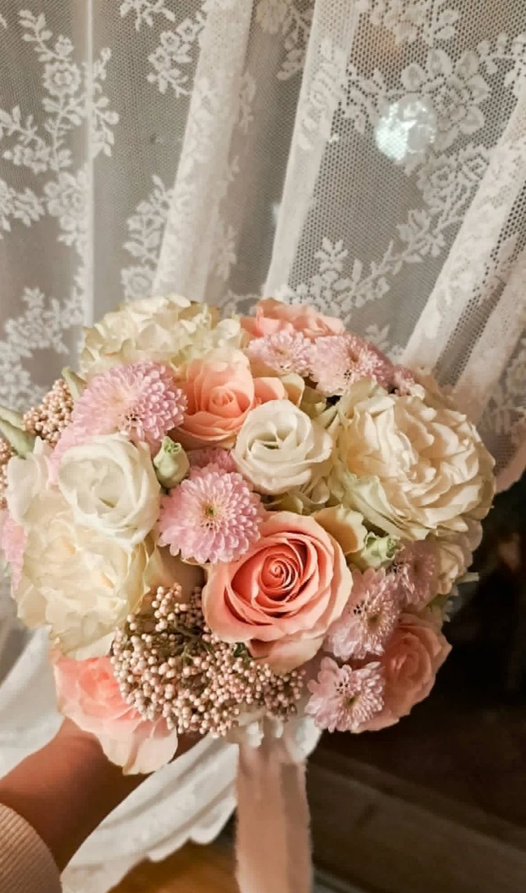 Bride holding romantic wedding bouquet of white roses, pink carnations, and coral roses with babys breath and white silk ribbon against lace curtain background