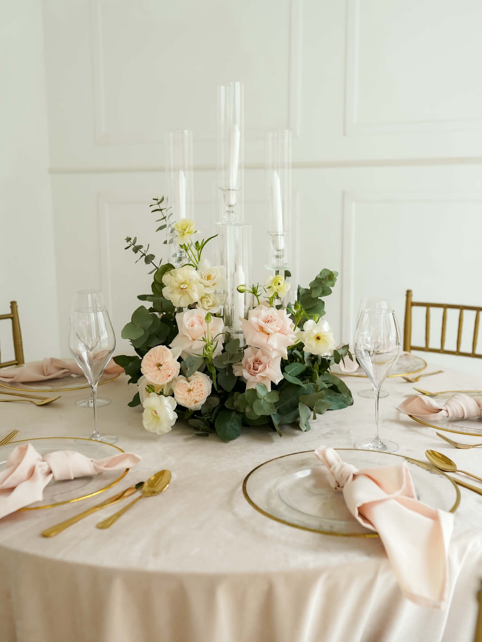 Elegant table setting with floral centerpiece of pale pink and white roses, tall white candles, gold cutlery, and blush napkins on round white tablecloth