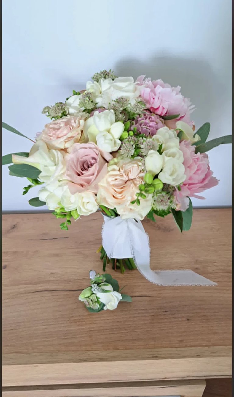 Bridal bouquet with white and pink roses, peonies, and greenery tied with a white ribbon on a wooden surface