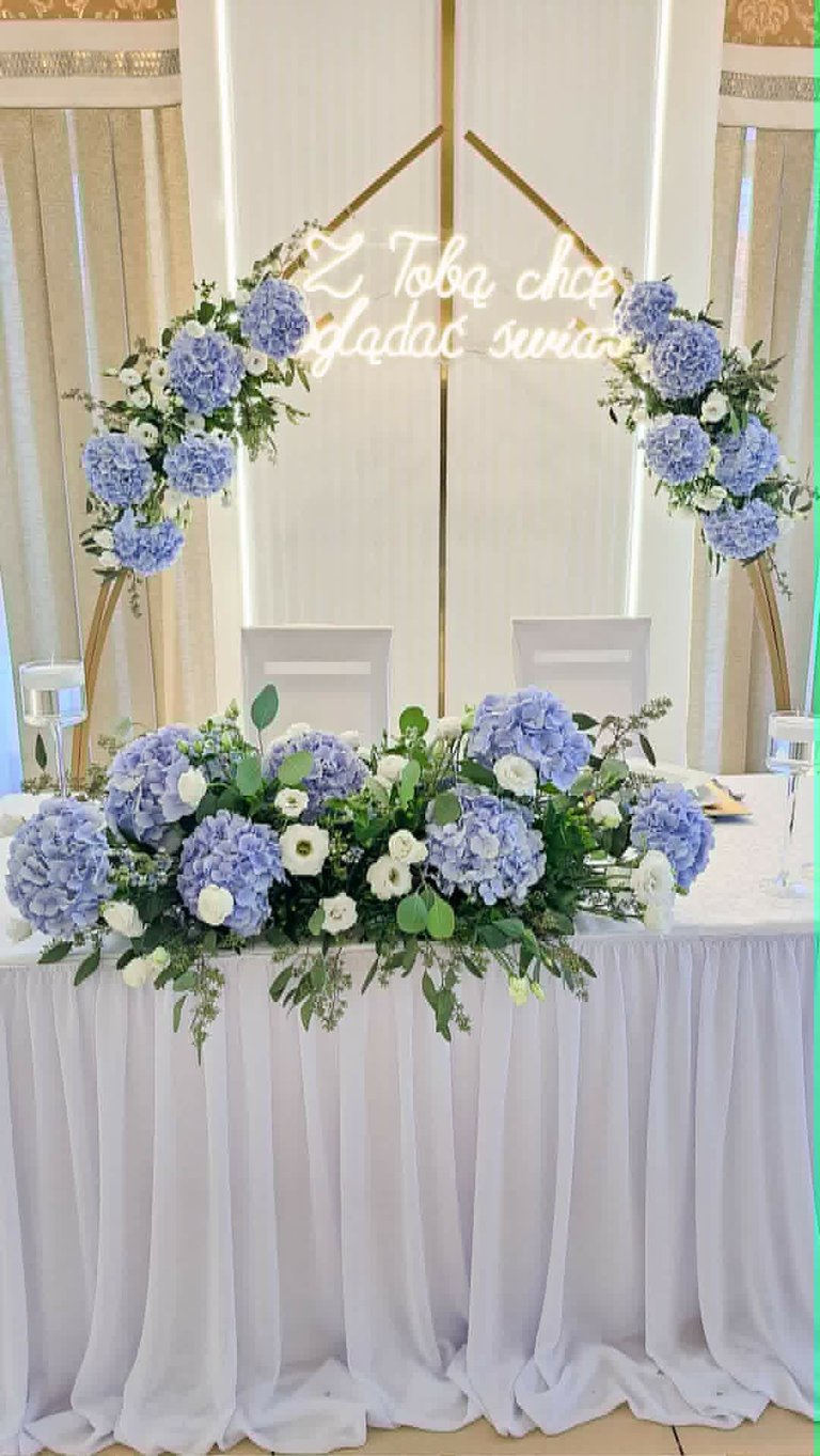 Elegant wedding sweetheart table with blue hydrangea and white flower arrangements, neon sign, and wooden branch arch