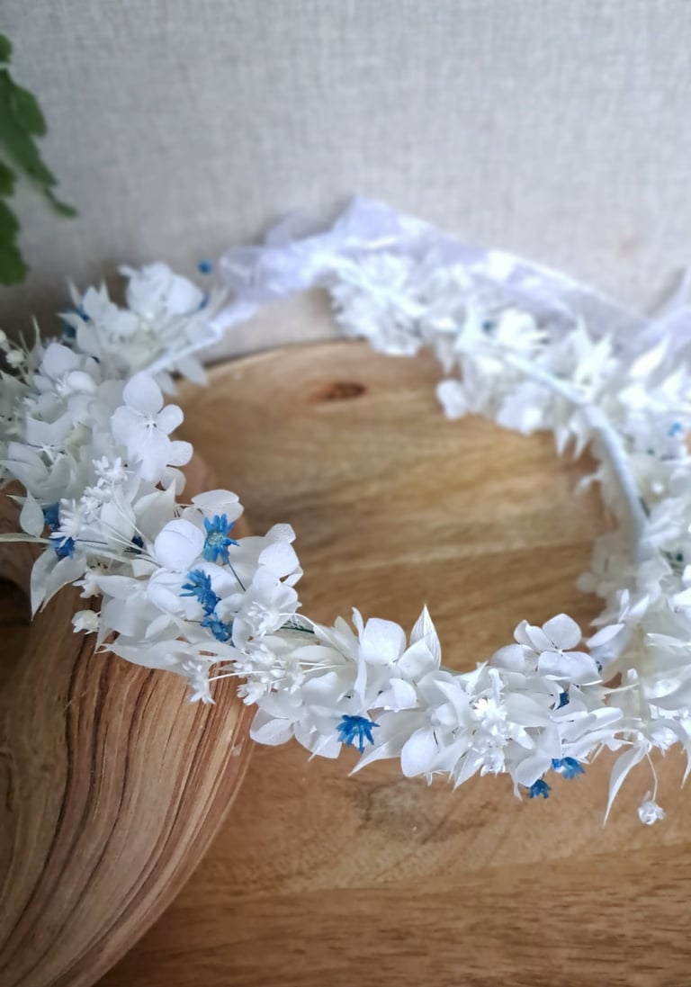 White flower crown with blue accents resting on wooden surface next to blonde hair and green foliage